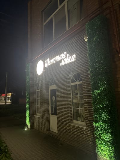 The image shows the exterior of a building at night in Yegorlykskaya, Russia. The building has a brick facade and is adorned with a horizontal strip of artificial greenery along the top. A prominent sign with white, illuminated Cyrillic lettering is mounted on the brick wall. The text on the sign reads "Цветочная лавка," which translates to "Flower shop."

Below the sign, a round, illuminated light fixture hangs from the ceiling of an overhang. To the right of the sign, there are several windows. One is an arched window with a grid pattern, and another is a rectangular window with a white frame. A small, dark sign with text is visible above the rectangular window.

A security camera is mounted on the building, near the greenery. The ground in front of the building appears to be paved with bricks or stones, and there are several small, upward-facing lights illuminating the greenery. In the distance, across a dark area that might be a street or a path, other buildings and streetlights are faintly visible. The overall atmosphere is calm and well-lit despite being nighttime. There are no people visible in the image.