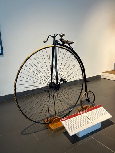 This media file shows a vintage "penny-farthing" bicycle displayed in a museum. The bicycle, also referred to as a "spider" bicycle in Russian, features a prominently large front wheel and a much smaller rear wheel. It is black with a rope-like material around the rim of the front wheel and has a bell and handlebars at the front. The bicycle is mounted on a wooden stand, casting a shadow on the tiled floor. To the right of the bicycle is a display board with text. The text, written in both Russian and English, describes the history of the penny-farthing bicycle, noting its popularity in the 1870s and its characteristics, including its dangerous nature due to a high center of gravity and the inconvenience of mounting and dismounting. It also mentions the notable 1884-1886 round-the-world bicycle trip by Englishman Thomas Stevens on such a bicycle. The background consists of a plain white wall with a subtle shadow. The lighting suggests an indoor setting, possibly a museum or exhibition space. The overall scene is static, focusing on the historical artifact and its accompanying information.