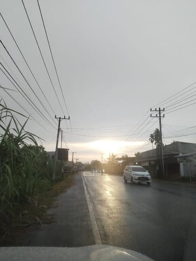 The image captures a serene, overcast morning or evening scene on a wet road in Putoh Sa, Indonesia. The sky above is largely covered in pale grey clouds, with a bright, soft golden light emanating from the low-lying sun near the horizon, slightly to the right of the center. This light casts a strong reflection across the wet asphalt road, creating a glistening path.
Numerous utility poles with intricate networks of power lines stretch across the frame, dominating the upper left and right portions of the sky. Tall, lush green vegetation, resembling reeds or sugarcane, borders the left side of the road, obscuring the ground beyond it. On the right, various simple, low-rise buildings and structures are visible, interspersed with a few palm trees and other foliage.
In the mid-ground, a white car, possibly a minivan or SUV, travels from right to left on the road, its front facing the viewer. Its headlights are off, but the vehicle is clearly illuminated by the sun's reflection. Further down the road, additional vehicle headlights are visible in the distance, indicating ongoing traffic. The foreground shows a partial view of what appears to be the hood of a vehicle from which the picture was taken, its surface a dull grey. No discernable text is visible on any signs or objects within the image. The overall impression is one of quiet transit through a semi-urban or rural area, marked by the gentle glow of the sun on a damp landscape.