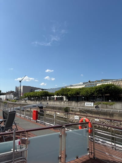 A long-shot, eye-level, outdoor daytime photograph shows a view from a boat's deck looking towards the Mainz waterfront. The deck features a railing with clear glass panels and a wooden top. In the foreground, a red and gray picnic table with two gray chairs is visible, along with a partially visible red fire extinguisher and a water bottle on a small metal shelf.
Beyond the deck, a concrete embankment lines a canal or river. A row of mature trees with full green foliage separates the embankment from a modern, multi-story building with a glass facade and light-colored awnings. A sign on the embankment reads "MAINZ".
In the distance, a construction crane is visible against a bright blue sky with scattered white clouds. The architecture suggests an urban setting in Germany, specifically Mainz, given the prominent signage. A few people are visible walking along the embankment. The water in the canal appears dark, with rocky edges along the embankment.