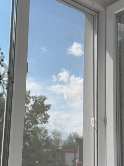 The image shows a view through a white framed window with a mosquito screen. The sky is blue with scattered white clouds. Green trees and the tops of buildings are visible in the background. The window appears to be open, with a white handle visible on the right side. The scene is likely from a residential building in Kyzyl, Russia, given the context of the request, but this cannot be definitively confirmed from the image alone. There are no people or specific actions depicted.