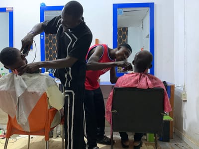 In a simple barbershop, likely in Gbeleko, Nigeria, two barbers give haircuts to their customers. One barber, wearing a black tracksuit with white stripes, uses an electric clipper on a seated customer draped in a white and gray barber cape, seated on an orange plastic chair. The second barber, clad in a red basketball jersey, focuses intently on the back of the head of a customer in a black office chair covered in a pink and red cape. Blue-framed mirrors are mounted on the white walls. The floor shows signs of wear and is speckled with debris. An electrical outlet is visible on the wall.