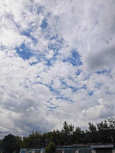 The image captures a cloudy sky over a collection of low-rise buildings and trees. In the distance, a tall, narrow structure with red and white lights is visible, possibly a communication tower or a small industrial chimney. The buildings appear to be garages or workshops, with corrugated metal roofs and brick and concrete walls. Green trees line the area, obscuring some of the structures and suggesting an urban or semi-urban setting. The clouds are a mix of white and grey, indicating overcast conditions, and patches of blue sky are visible between them. The scene is devoid of people or any apparent activity.