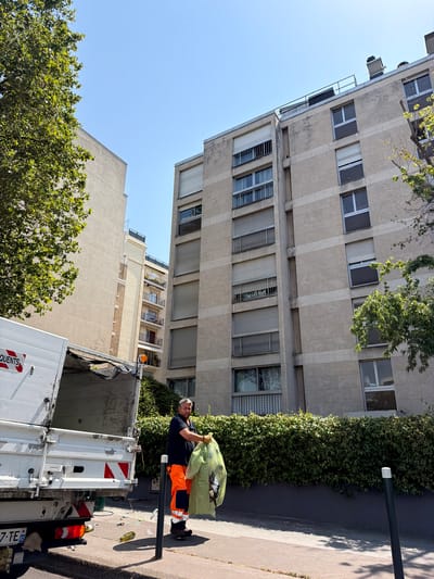 A man, wearing an orange work uniform, stands on a sidewalk next to a white garbage truck, holding a yellow plastic bag full of trash. The scene appears to be in a residential area in Neuilly-sur-Seine, France, as suggested by the apartment buildings and trees in the background. The sun is shining, indicating it is daytime, and the weather seems pleasant. The man is looking at the camera and has a neutral expression. The garbage truck is parked, and its back doors are open, suggesting it is in the process of collecting trash. The sidewalk is paved, and there are some loose pieces of trash scattered on the ground. One of the apartment buildings has a vine growing up its facade. The garbage truck has some text on its side, "QUENTS," which is partially visible. The license plate of the truck is visible and reads "7-TE S2".