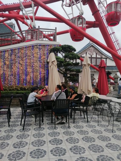 The scene depicts an outdoor café setting in Hong Gai Ward, Vietnam, situated at the base of a large red Ferris wheel. The cafe area is paved with grey and white tiles in a floral pattern, with black metal tables and chairs arranged for several groups of people.
A group of individuals are seated at one of the tables, seemingly engaged in conversation. Other individuals are seated at nearby tables, some facing away from the camera. A few unopened beige umbrellas and a red umbrella stand near the tables.
In the background, a stone wall adorned with strings of small red, white, and blue lights provides a backdrop. A traditionally styled white and brown building with a dark tiled roof is visible behind the wall. The red Ferris wheel dominates the scene, with its cars numbered "18". The sky appears overcast, suggesting a cloudy day.
