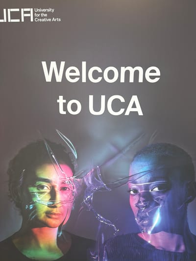 The image shows a poster with text and two portraits. The text at the top left reads "UCA" in a stylized font, with "University for the Creative Arts" written below it. Below this, larger white text states "Welcome to UCA". The bottom half of the poster features two portraits of individuals with artistic lighting and effects. The person on the left has colorful lights cast on their face, with what appears to be a transparent, fragmented material overlaying their features. The person on the right also has dramatic lighting in shades of blue and purple, with similar fragmented material over their face, highlighting facial features. The background is a dark, textured gray. The poster is likely displayed at the University for the Creative Arts campus in Epsom and Ewell, United Kingdom.