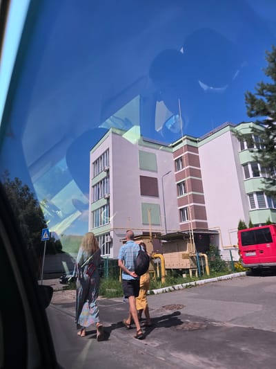 This media file depicts a street scene in Petropavlivska Borshchahivka, Ukraine, as seen through a car windshield, implying a reflection. The image is oriented sideways.
The main subjects are two people walking on a paved sidewalk next to a building. One person, wearing a plaid shirt and dark shorts, carries a backpack. The other person is wearing a colorful, long dress. They appear to be walking in the same direction. Shadows suggest it is daytime, likely afternoon, under clear, sunny weather.
The setting includes a modern-looking building with green and brown accents and numerous windows. Yellow pipes are visible on the side of the building, suggesting some kind of utility infrastructure. A red bus is parked to the right of the building. The ground is asphalt, and there is some greenery and trees in the background and along the sidewalk. A blue pedestrian crossing sign with a white pictogram is also visible on the sidewalk.
The reflections on the windshield create a sense of depth and overlay the scene with the blue sky and some tree foliage. The overall impression is a snapshot of everyday life on a sunny day in a Ukrainian town. No discernible text is visible in the image.