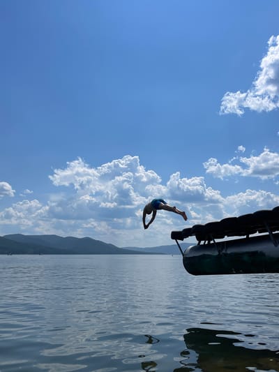 The image depicts a person diving into a body of water, likely Krasnoyarskoye Reservoir near Divnogorsk, Russia. The sky is blue with scattered white clouds. In the foreground, the edge of a pontoon boat or dock is visible, adorned with black tires for protection. A person, wearing blue swim trunks, is captured mid-air, executing a dive with their body arched and arms extended. The water is calm with ripples reflecting the sky and surrounding landscape. In the distance, rolling green hills or mountains form the horizon. Two small boats are visible on the water, adding to the sense of scale. The overall scene captures a moment of recreation on a sunny day.