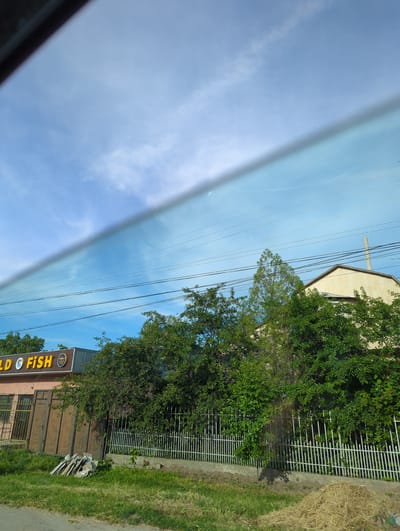 The image, taken from inside a vehicle, shows a view of Beş-Küŋgöy, Kyrgyzstan. The right side of the image contains a building with yellow "GOLD FISH" signage. There's a white fence, trees, and green grass alongside the building. Overhead are power lines against a blue sky, partly obscured by a car window.
