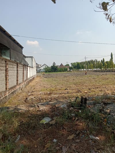 The image shows an outdoor scene with a clear blue sky and a dry, grassy field. In the foreground, there is a pile of dried leaves, grass, and some litter, including plastic bags. A brick wall, part of a building, runs along the left side of the image. The wall is constructed with grey concrete blocks and has red brick trim. Beyond the wall, there are some buildings with white facades and terracotta-tiled roofs. In the middle ground of the field, a person wearing an orange shirt and a hat is visible, seemingly working or walking. Further in the background, a line of tall, slender green trees borders the field. Power lines and poles are also present in the distance. The overall impression is of a rural or semi-rural area undergoing some form of development or preparation, possibly agricultural land. The specific location is given as Kemamang, Indonesia.
