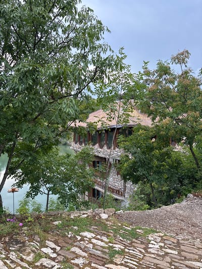 This detailed outdoor photograph, taken in Lukovit, Bulgaria, presents a multi-story building positioned beside a body of light green water, viewed through dense deciduous tree foliage from an elevated perspective. The foreground consists of an uneven path paved with rough, light-colored stones interspersed with green grass and small weeds. A cluster of small purple flowers is visible on the left, near a thin metal stake topped with a decorative, flower-shaped metallic object. This path transitions into a coarser, gravelly ground surface towards the right.
The building, partially obscured by trees, features a sturdy lower section constructed from irregular grey and brown stone blocks. Its upper visible level includes a series of distinctive, curved windows with dark wooden frames, offering expansive views of the adjacent water. The roof is complex, tiled with weathered reddish-brown and orange tiles, and appears to have multiple gables or dormer structures. The calm water, a pale green or turquoise hue, reflects the surrounding environment. Various trees with vibrant green leaves, some exhibiting distinctive seed pods, frame the scene, with one prominent tree on the left foreground. The sky above is uniformly overcast and light blue-grey. No human figures are present in the image.