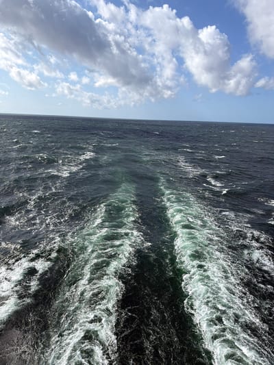 The image depicts a turbulent wake left by a vessel moving through dark, choppy water under a partly cloudy sky. The water's surface is a deep blue-green, with white foam and spray churned up by the passage of the boat. The wake extends away from the viewer, forming two distinct, frothy trails that gradually dissipate into the open sea. The horizon line is visible, separating the water from the sky, which is dominated by large, puffy white and grey clouds against a bright blue background. The scene suggests a sense of motion and the vastness of the ocean. The location is not specified as being in the city of Odargowo, Poland; the image solely shows an open body of water. There are no people, subjects, objects other than the water and sky, or actions depicted in the image that would indicate a specific urban location.