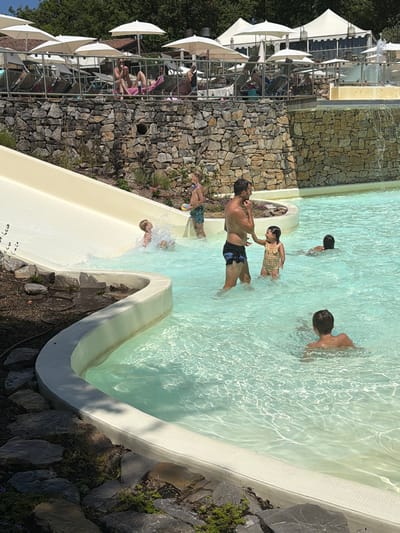 This image depicts a bright, sunny day at an outdoor swimming pool, likely a resort or water park, situated in the vicinity of Cavriglia, Italy. The foreground features a light-colored water slide curving into a shallow, clear turquoise pool. Several people are visible interacting in and around the water.
In the pool, a man in dark swim trunks stands partially submerged, looking towards a young girl in a checkered swimsuit who is playfully reaching for his hand. Further back in the pool, another young boy is swimming. Near the bottom of the water slide, a child is splashing as they emerge from the slide into the water, while another child stands at the top of the slide's entry.
The background reveals a stone wall constructed from various-sized rocks, supporting a raised area with lounge chairs and umbrellas. Beyond this, buildings with white tent-like structures and more umbrellas are visible, suggesting amenities for relaxation. Lush green trees form a backdrop against a clear blue sky. The overall scene conveys a leisurely atmosphere associated with a vacation or recreational outing.