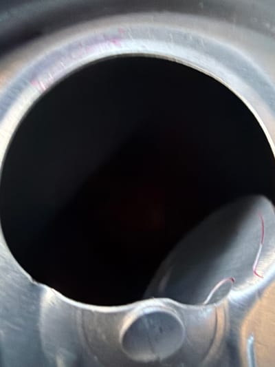 This is a close-up, overhead view of the interior of an open metal can. The dominant features are a large, dark, circular opening, likely the mouth of the can, which takes up the majority of the frame. The interior of the can appears to be dark, possibly reflecting a dark liquid or shadow. Surrounding the opening is the metallic rim of the can, which shows some subtle color variations, including faint pinkish or purplish streaks. To the right of the opening, a curved section of the can's interior wall is visible, with a small, flat piece of what appears to be plastic or metal detached and resting on the surface. This detached piece has a thin red thread or fiber adhered to it. The overall impression is of a utilitarian object, possibly a food or beverage container. There is no indication of the city of Kareby, Sweden, or any environmental context.