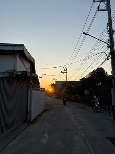 This image depicts a street scene at sunset within Chiang Rai City Municipality, Thailand. A paved two-lane road extends into the distance, illuminated by the low sun positioned directly ahead, casting long shadows. The sky transitions from a bright orange-yellow at the horizon to a pale blue overhead.
On the left, a two-story concrete building with louvered vents is partially visible behind a tall, dark metal gate and a light-colored concrete wall. The right side of the street features multiple utility poles with dense bundles of overhead electrical and communication wires; a prominent pole in the foreground includes a streetlight fixture. Further down the road on the right, foliage and additional structures are visible.
Two individuals on motorcycles are present. In the mid-ground, a dark-colored motorcycle with an illuminated headlight is moving towards the viewer; the rider is silhouetted. In the foreground on the right, a light-colored scooter-style motorcycle is stationary, positioned near a dark metal railing. Its rider, wearing a light-colored helmet, is seated on the vehicle.