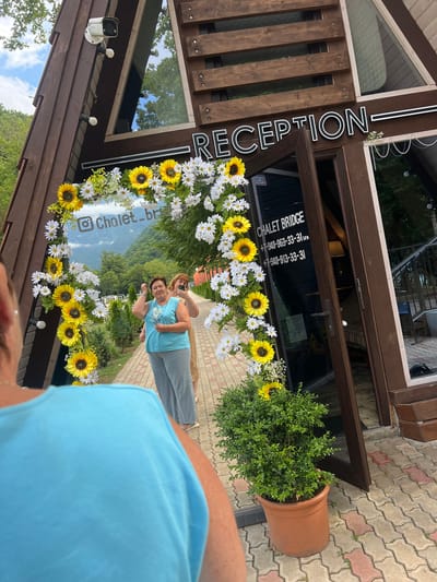 The image shows two women standing in front of a building labeled "RECEPTION." They are framed by a floral archway made of sunflowers and daisies. The building appears to be a lodge or hotel, with wooden siding and a glass entrance. The setting is outdoors, with a view of lush green hills and a cloudy sky in the background, suggesting a daytime scene.
One woman, wearing a blue tank top and grey pants, is smiling and looking towards the camera. The other woman, behind her, is wearing a beige top and also appears to be looking at the camera, possibly taking a photo. The overall mood seems relaxed and cheerful.
Text visible in the image includes "RECEPTION" on a vertical sign. On the glass door, there is text that reads "CHALET BRIDGE" followed by phone numbers: "+7-940-963-33-31 SPA" and "+7-940-913-33-31". A social media handle "@chalet_bridge" is also visible on a circular element within the floral arch. A security camera is mounted on the building's exterior. The ground is paved with interlocking bricks.