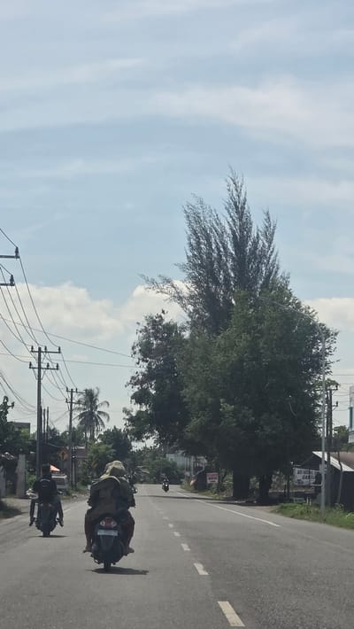 A slightly tilted, full-day shot shows a straight asphalt road in Blang Lilue, Indonesia. Power lines and trees are on either side. Two motorcycles are driving towards the camera. The sky is partly cloudy. A sign in Indonesian is visible.