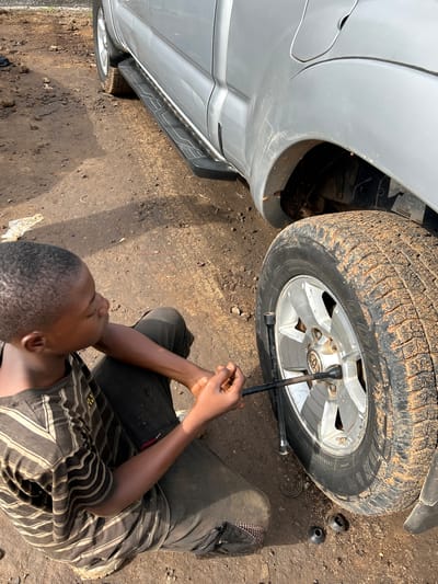 A young Black boy, wearing a brown and tan striped t-shirt and dark plaid pants, kneels on the dirt ground in Barikin Jirga Summa, Nigeria, working on the rear passenger-side tire of a silver pickup truck. The tire is caked in mud. The boy is using a lug wrench to loosen the lug nuts on the wheel. The truck is parked on a dirt road, with debris and loose soil scattered around. The sun casts shadows on the ground.