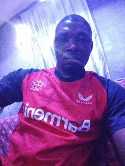 A dark-skinned person is centered, wearing a red Bayer Leverkusen soccer jersey with "Barmenia" in white lettering and the Castore logo. The jersey has gray sleeves. The person is seated indoors, on a pink paisley patterned cushion. Behind is a white sheer curtain, partially lit by a bright light source. The overall image is slightly blurry and washed in blue light.