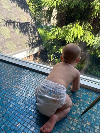A baby wearing a diaper lies on their stomach on a blue tiled surface near a window. The baby is looking out the window at a green, leafy tree. Outside the window, the ground is covered in patterned tiles with shadows from the tree. A table leg is also visible on the tiled surface. The location of the image is Чёрное, Russia.