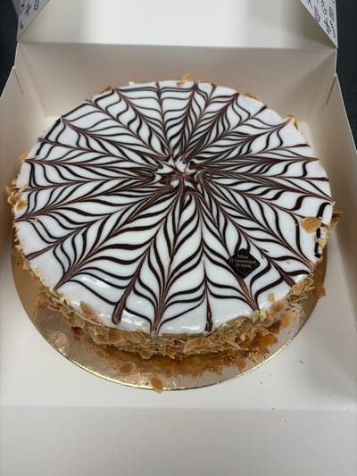 A circular cake with white icing and a chocolate spiderweb design is presented in a white cardboard box. The cake is decorated with a complex pattern of brown chocolate lines radiating from the center, creating a spiderweb effect. The sides of the cake are coated with slivered almonds. A small, dark square plaque with text is visible on the top surface of the cake. The cake is placed on a golden cardboard base. The box appears to be a bakery box, with decorative patterns on the interior flaps, suggesting it was purchased from a bakery in Hœrdt, France, as indicated by the text on the plaque. The background is out of focus, showing the interior of the white box and a dark surface beyond.