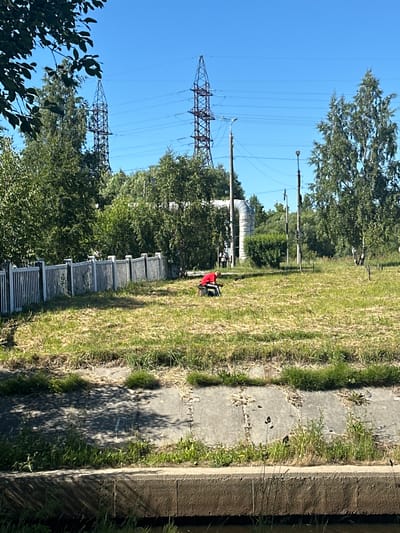 The image depicts an outdoor scene with a person in a red shirt and black pants crouching in a grassy area. The person is seated on a dark-colored stool or low object. In the background, there is a white fence, trees, power lines supported by tall metal towers, and a large industrial pipe. The area appears to be a park or undeveloped land, possibly near an industrial zone, given the presence of the power lines and pipe. The weather is clear with a blue sky. Based on the context of the provided information, this scene is located in the city of Severodvinsk, Russia.