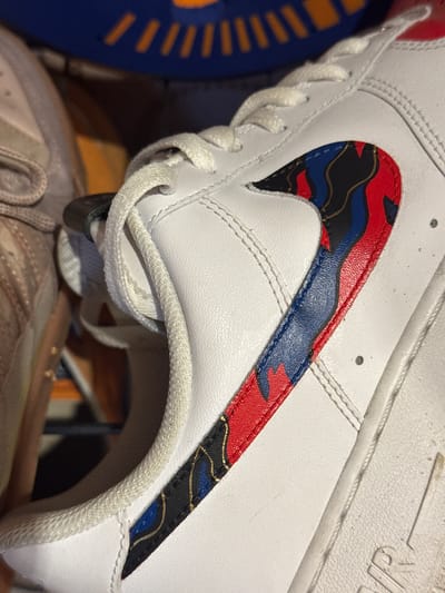 A close-up image shows the side of a white leather sneaker. The iconic "swoosh" logo is present on the side, but it is filled with a complex, patterned design in red, blue, black and gold. The laces of the shoe are white and knotted. Portions of other objects (likely other shoes) are visible in the background, with one appearing blue and another a lighter, brownish-grey. The lighting suggests indoor conditions. The location is not determinable from the image.