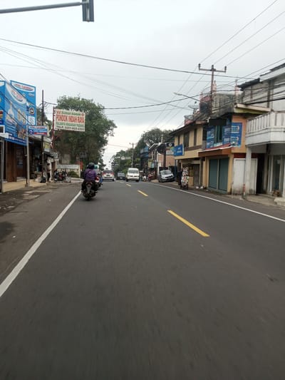 The image captures a street scene with a two-lane road marked by a solid white line on the left and a dashed yellow line in the center. A person on a motorbike is traveling along the road, with other vehicles visible in the distance. Buildings line both sides of the street, with signs and advertisements visible on some of them. Power lines stretch across the sky. The sky is overcast. Signage indicates "INDO LAUNDRY" and "RUMAH MAKAN PONDOK INDAH RAYA, RAJANYA MASAKAN PADANG TELP. 0251-825-2323."