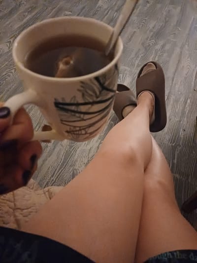 This is a close-up, high-angle shot of a person's legs and a mug of coffee. The person is wearing brown slippers and dark shorts. They are sitting on what appears to be a textured, light-colored surface, possibly a rug or blanket. The floor, visible in the background, is made of wood-look planks with a greyish-white tone and a wood grain pattern.
The primary object in focus is a ceramic mug, held in a person's hand, containing a dark liquid, presumably coffee. The mug has a distinctive pattern of black, vertical, irregular lines. A spoon is visible inside the mug. The person's hand, with painted nails, is also visible. The lighting suggests an indoor setting, possibly in the evening or on a cloudy day. The overall mood is relaxed and casual. There is no visible text in the image. The location context provided is Trypillia, Ukraine.