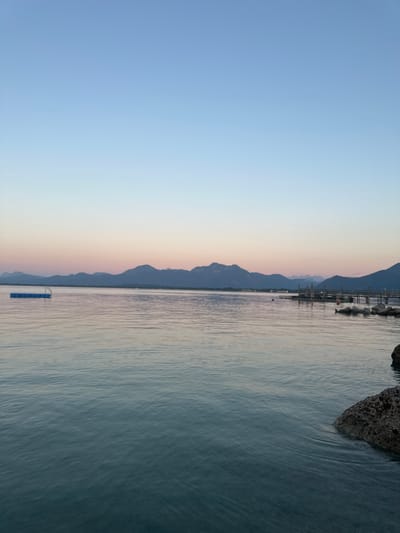 An expansive, calm body of water, likely Lake Chiemsee, dominates the foreground and middle ground. The water reflects the soft hues of a twilight sky, transitioning from a pale blue at the top to gentle pinks and oranges near the horizon. In the distance, a range of dark, silhouetted mountains forms a dramatic backdrop.
To the left of the frame, a blue floating platform with a white ladder is anchored in the water. Further to the right, a wooden pier extends out from the shore, with several structures and what appear to be boats moored alongside. The shoreline is marked by a few dark rocks in the lower right corner of the image, with one prominent rock emerging from the water.
The scene suggests a peaceful evening in Prien am Chiemsee, Germany, a town known for its picturesque location on the lake and its proximity to the Bavarian Alps. The clear sky and still water indicate favorable weather conditions. There are no people visible in the image.
