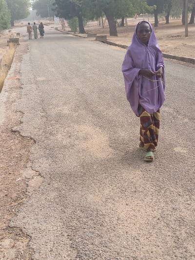 The image portrays an outdoor scene in Kanti, Nigeria, focusing on a young individual walking on a paved road. The individual, positioned in the right foreground, wears a light purple hooded garment, patterned trousers featuring geometric shapes in brown, yellow, and red, and light green sandals. They hold several thin, light-colored strands in their hands and look directly forward. The asphalt road, showing signs of wear and cracking, extends into the background, where three additional figures are observed walking away from the viewer. The road is bordered by dry verges with exposed concrete curbing. Numerous deciduous trees with visible trunks and sparse foliage line the right side of the road and extend into the distant background, indicating a dry, possibly semi-arid, environment. The scene is illuminated by natural daylight.