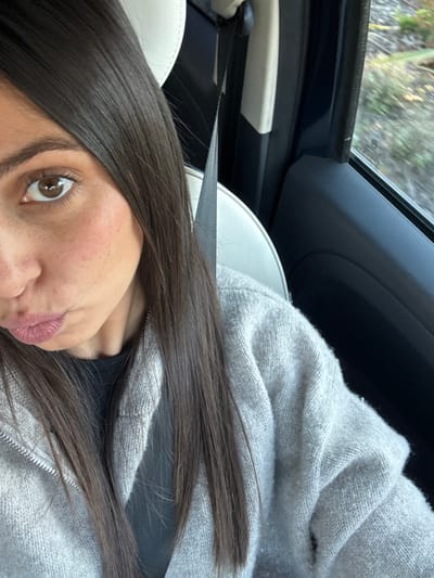 A young adult female is depicted from the chest up, seated in the passenger side of a vehicle, likely in Antibes, France. She wears a light gray knit zippered cardigan over a dark-colored top and is secured by a black seatbelt that crosses diagonally over her chest. Her long, straight, dark brown hair falls forward over her shoulders. She looks directly towards the viewer with brown eyes, and her lips are pursed into a pout. The white headrest of the vehicle seat is visible behind her. Through the vehicle's window on the right, blurred green and brown vegetation is visible, indicating the vehicle is in motion along a roadside environment.