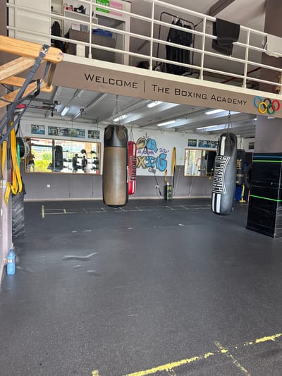 This is a photograph of the interior of a boxing gym in Calp, Spain. The gym features several punching bags hanging from the ceiling, one of which is a large, dark-colored bag with white lettering that reads "CHARLIE." Another, lighter-colored punching bag is suspended above it. The walls are decorated with posters and graffiti, including a stylized depiction of a boxing glove and the words "LA ACADEMIA BOXING CLUB." A sign on the left side of the frame reads "WELCOME | THE BOXING ACADEMY." In the background, there are shelves filled with various items, including what appear to be gym equipment and clothing. A mirror is visible on the back wall, reflecting the gym space. The floor is dark, likely rubber or a similar material common in gyms, with yellow markings. The lighting suggests it is daytime, with artificial lights also visible. The overall impression is of a functional and active training facility.