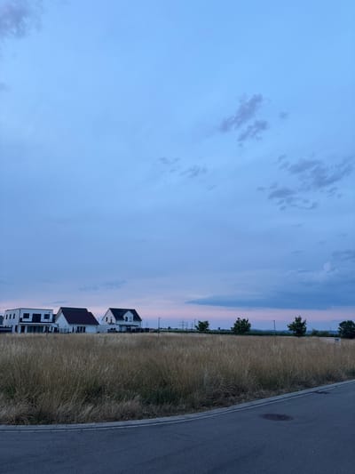 The image depicts a rural-urban fringe area in Bönnigheim, Germany, at dusk. In the foreground, a paved road curves from the bottom right corner towards the left, with a concrete curb bordering a field of dry, tall grass. Beyond the grassy field, three modern houses are visible. The leftmost house is a two-story structure with a white facade and dark accents, featuring balconies. The middle house is a white, two-story building with a dark, pitched roof. The rightmost house is also a two-story white structure with a dark, pitched roof and dark window frames. In the background, a line of trees and utility poles stretch across the horizon. The sky is a gradient of deep blue at the top, transitioning to lighter blues and then hints of pink and purple near the horizon, indicating the twilight hour. There are scattered clouds throughout the sky. The scene is devoid of people or any discernible activity.