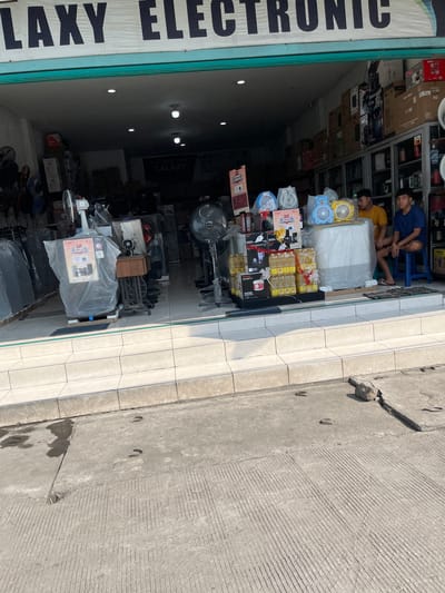 The image shows the exterior of an electronics store in Gedang, Indonesia. The store is named "GALAXY ELECTRONIC" and is filled with various electronic appliances, including fans, refrigerators, and other household items. Two young men are sitting inside the store, one wearing a yellow t-shirt and the other a blue t-shirt. The store appears to be located on a street with a concrete sidewalk and road. The storefront is open, revealing the merchandise displayed inside. Several fans are visible, including a large standing fan, a desk fan, and two smaller decorative fans. Bottles of cooking oil are stacked on shelves, along with other packaged goods. The overall impression is of a busy retail environment in an Indonesian city.