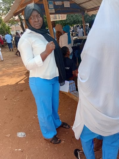 A woman in a hijab, white blouse, and blue pants is shown in a medium shot. She looks at the camera. A white cloth is draped above her, partially obscuring other seated figures. Other people sit behind her. The setting appears to be outdoors, with a dirt ground and a building in the background. The photo seems to be taken at a slight angle.
