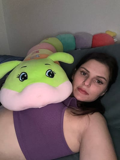 A woman is lying down with a large stuffed caterpillar toy. The toy is lime green with pink accents and has large cartoon eyes. The woman is wearing a purple ribbed tank top. The background appears to be indoors, possibly a bedroom, with a wall and furniture. The lighting is dim. The image does not provide any information about the city of Novoselye, Russia.
