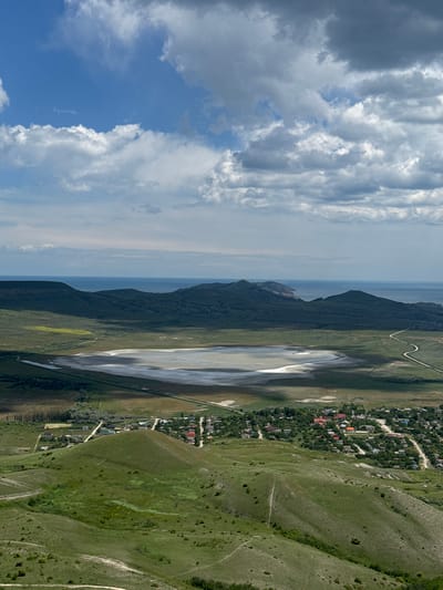 An aerial shot of Коктебельский поселковый совет, Ukraine, shows a landscape where mountains meet the sky. The left portion of the image features a sky with cumulus clouds, moving towards the horizon. A sliver of ocean is discernible along the horizon under light cloud cover. A line of mountains runs along the middle of the image. To the right of the mountains, there is a large lake. Adjacent to the lake is a town. Rolling green hills with paths are on the right side.