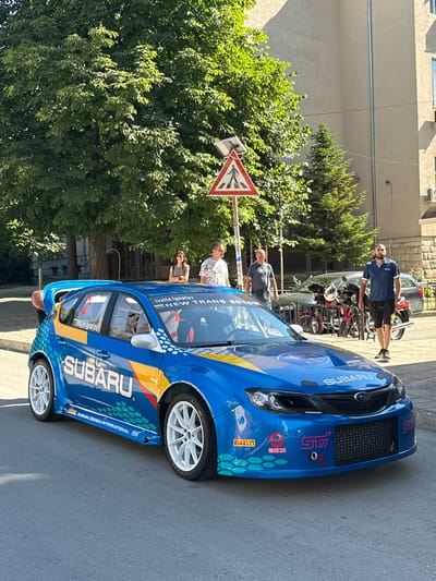 The image shows a bright blue Subaru Impreza WRX STI rally car parked on an asphalt street in Gorna Oryahovitsa, Bulgaria. The car is highly decorated with sponsor logos, including "SUBARU" prominently displayed on the side and hood, "STi" on the front bumper, and smaller logos for Pirelli and Sparco. The car has white racing wheels and a large rear spoiler.
Behind the car, several people are visible. On the left, a man is leaning into the driver's side of the car. Further back, a woman stands looking towards the car. To her right, two men are standing near a pedestrian crossing sign. In the background, there is a building with beige siding and a dark-colored sedan and motorcycles. Trees line the street, and the sky is clear and blue. The overall scene suggests an outdoor event or car display.