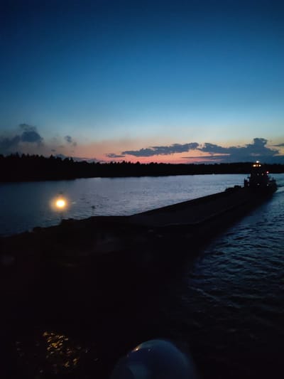The image depicts a dark, wide river at dusk. A long, dark barge is visible in the foreground, illuminated by a single light at its front. A towboat pushes the barge upriver. A light on the shore reflects off the water. A silhouetted treeline is visible on the distant shore. The sky transitions from dark blue to a band of orange and pink at the horizon, with clouds present. The location in the city of Нова, Russia, is not visually verifiable from the image.