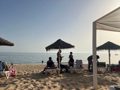 A wide-angle exterior view captures a beach scene at Praia do Almancil, Almancil, Portugal, under clear daylight conditions with the sun low on the horizon. The foreground consists of light golden-brown sand with visible undulations and footprints.

From left to right:
On the far left, a portion of a thatched beach umbrella is visible above two light-colored sun loungers, one draped with a pink and white striped towel.
In the mid-left, a second thatched beach umbrella shades three individuals. One person is seated on a sun lounger, facing the ocean, wearing a dark top. Two other individuals stand nearby; one, largely obscured, and another wearing a dark top and patterned shorts, holding an object near their waist.

The calm, light blue-green ocean extends across the mid-ground, meeting a clear, light blue sky at the horizon. A lone individual, silhouetted, stands waist-deep in the shallow water in the center of the frame, facing away from the camera. Two small boats are visible on the distant horizon.

The right side of the frame is dominated by a large white fabric canopy structure with a prominent white support pillar in the immediate foreground. Beneath this canopy, a third thatched beach umbrella is positioned. Two individuals are partially obscured by the canopy structure: one person with dark, possibly braided hair is bending over near a sun lounger, and another is seated on a sun lounger. Beach towels and bags are placed on or near the loungers.