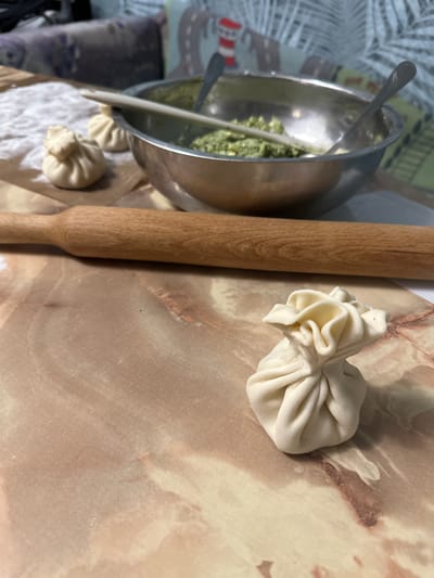 The image shows a culinary setup, likely for making dumplings or a similar dish. A stainless steel bowl containing a green filling, possibly spinach or herbs, sits to the left. A wooden rolling pin lies horizontally next to the bowl. On a beige surface with a marble-like pattern, there's a partially-made dumpling, pinched at the top to form a bundle. Another similar dumpling is visible in the lower left corner. The image does not provide evidence to support the location being Pokrov, Russia.
