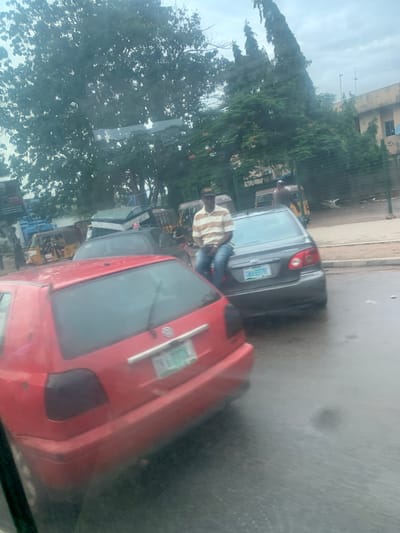 A red car is parked on a street in Aso, Nigeria. The car is a Volkswagen Golf MK4 and is facing left. The car is dirty and has some scratches on the body. There is a man sitting on the trunk of the car. The man is wearing a striped shirt and jeans. He is looking at the camera. There is a gray car parked behind the red car. The gray car is a Toyota Corolla. There are two people sitting in the gray car. They are looking at the camera. There are several other cars and a motorcycle parked on the street. The street is wet, indicating it has recently rained. The sky is cloudy. There are trees and buildings in the background. The scene appears to be a street in a residential area or a market.