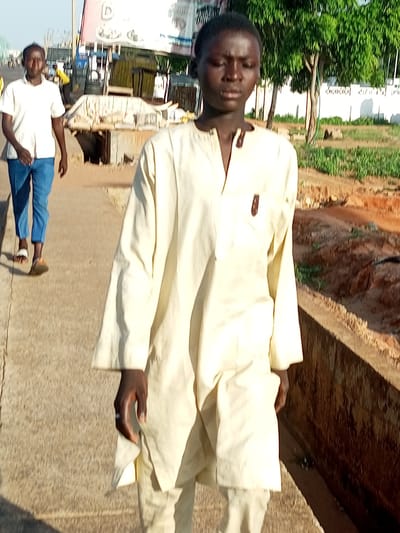 In Garko, Nigeria, two individuals walk along a paved path. The foreground is dominated by a person wearing a cream-colored robe and matching pants. Their gaze is directed downwards and slightly to the right. To the left and slightly behind, a second person is visible, wearing a white short-sleeved shirt and blue trousers. They are looking forward. The background shows a bustling environment with market stalls, trees, and buildings. A sign with the letter "D" is prominently displayed on a structure in the background. The ground appears to be a mixture of concrete and reddish-brown earth.