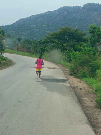 A lone male runner is captured from behind as he jogs along a paved road. The man is clad in a pink tank top and yellow shorts with blue accents. The road, slightly damp, curves gently to the left and is flanked by lush green vegetation. In the distance, a large, rocky hill dominates the landscape. This scene is in Kaninkon, Nigeria.