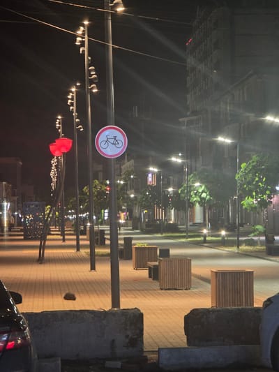 The image depicts an outdoor scene at night, likely on a street or promenade, with a focus on a row of streetlights and decorative elements. The lighting suggests it is after dusk. Several streetlights, some with multiple lamps, illuminate a paved walkway and an adjacent area with trees.

Prominently featured is a circular red and white sign with a bicycle symbol, indicating a bicycle path or restriction. Further down the path, a red, heart-shaped or petal-shaped light fixture is visible, adding a decorative touch. Rows of modern planters, made of wood, are arranged along the walkway. In the background, buildings with illuminated windows can be seen, suggesting an urban or town setting.

The overall atmosphere is calm and perhaps a bit serene, characteristic of an evening street scene. The paving stones and the design of the planters suggest a well-maintained public space. There is a sense of urban planning and design evident in the arrangement of elements. A portion of a car is visible on the right side of the image. The time of day is clearly night due to the artificial lighting and the darkness of the sky.
