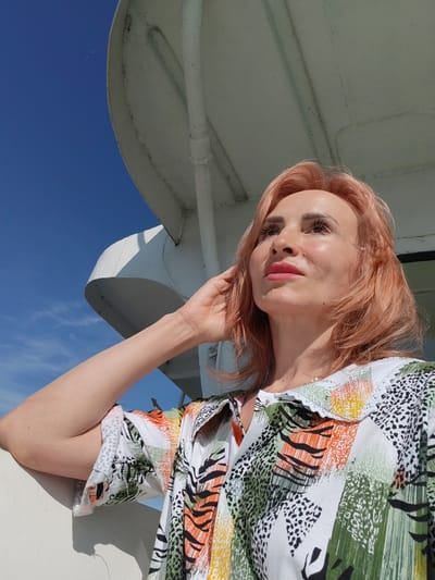 The image features a woman with short, peachy-pink hair, looking up and to her right with a contemplative expression. Her skin is illuminated by sunlight, and she is wearing a colorful, patterned shirt with a white collar. The pattern includes abstract shapes in green, orange, yellow, and black, reminiscent of animal prints and brushstrokes. She has her right hand raised, touching her hair or temple.
The setting appears to be outdoors, with a clear, bright blue sky visible in the background. Above and behind the woman, there is a large, white structure, possibly part of a boat or building, with pipes and curved elements. The sunlight suggests it is daytime, likely midday given the brightness. The woman's gaze and the bright lighting evoke a sense of peace or reflection. The context provided places the location in Tsikovo, Russia. No visible text is present in the image.