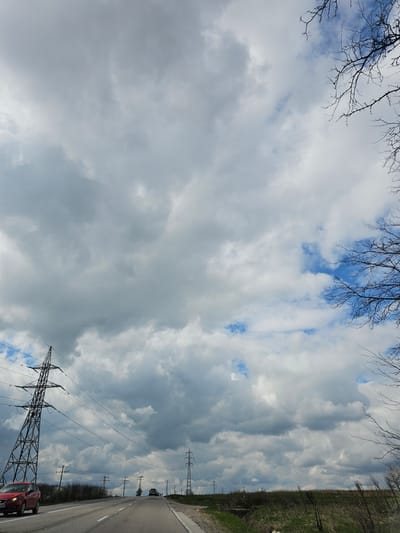 The image depicts a view from a road in Tvardintsi, Bulgaria, angled upwards towards the sky, which is filled with mostly grey and white clouds, with patches of blue sky visible. Bare tree branches are visible in the upper left corner of the frame. The road runs along the right side of the image, with grassy terrain and a sparse line of trees along the edge. Utility poles and power lines stretch along the right side of the frame, converging toward the horizon. A red car is parked on the roadside in the lower right corner. A tall metal electricity pylon is visible in the lower right.