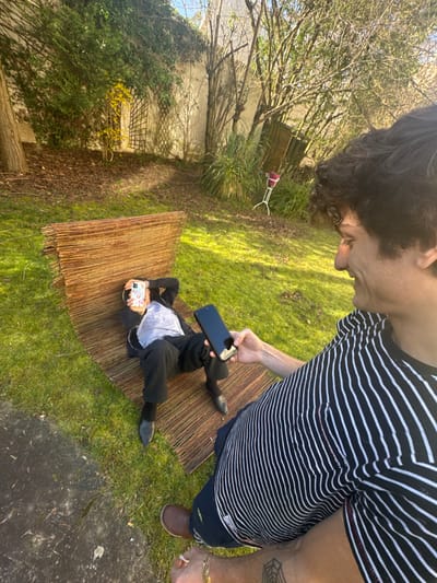The scene is set in a sunlit residential garden in Vincennes, France. In the foreground, a male with dark, curly hair, wearing a black and white striped t-shirt, dark shorts, and brown shoes, is positioned with his right side partially visible. His right arm is extended, holding a black smartphone horizontally, aimed towards another individual. A gold-colored chain is visible on his left wrist, and a spiderweb tattoo is present on his right forearm. He exhibits a slight smile.
In the mid-ground, a second male is lying supine on a reddish-brown woven mat, which appears to be made of bamboo or reeds and is laid out on green grass. This individual is dressed in a dark jacket or hoodie over a light-colored shirt, dark pants, and dark shoes. He holds a white-cased smartphone directly in front of his face, obscuring its details.
The background consists of a tall concrete wall extensively covered with green ivy and other foliage. Various trees and shrubs, some with sparse leaves and others bare, are visible. A white metal plant stand holding a magenta pot is situated on the grass in the upper-right background. The ground is primarily green grass, with patches of exposed earth and a segment of paved or concrete surface in the lower-left foreground. The area is illuminated by bright sunlight, creating visible shadows.