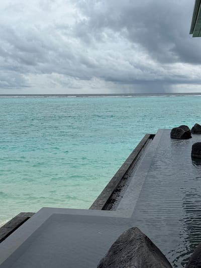 This image shows a view of the ocean from what appears to be an elevated structure or deck. The ocean water is a vibrant turquoise color and appears shallow with some ripples. In the foreground, there is a dark, angular object that seems to be part of a man-made structure, possibly made of concrete or stone, with some large, dark rocks placed on top. The water cascades over the edge of this structure, creating a subtle waterfall effect.

To the left of the frame, the sky is filled with dramatic, dark, and fluffy clouds, suggesting overcast weather or a storm approaching. A sliver of the ocean horizon is visible between the clouds and the water in the foreground. The overall mood is somewhat dramatic due to the contrast between the bright turquoise water and the dark, moody sky and foreground elements. The time of day is not clearly discernible, but the lighting suggests it could be daytime, perhaps late afternoon.

There is no visible text in the image. The location cues are limited to the appearance of the ocean and sky, which might suggest a tropical or coastal setting, but the specific location is unknown.